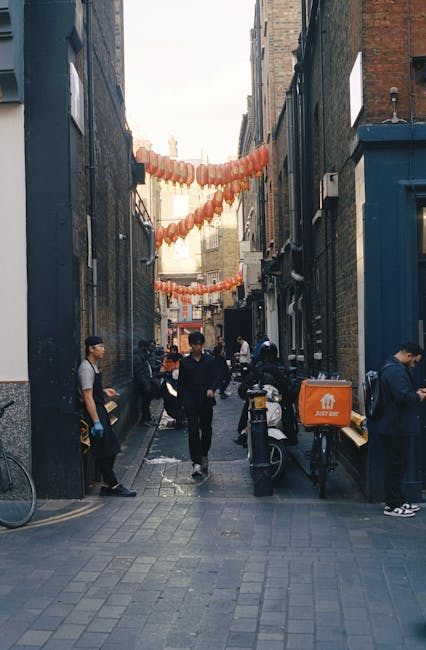 A narrow urban alleyway flanked by brick buildings with various signs, including one for a family development society, and a network of overhead wires. The ground is paved with concrete, featuring sma