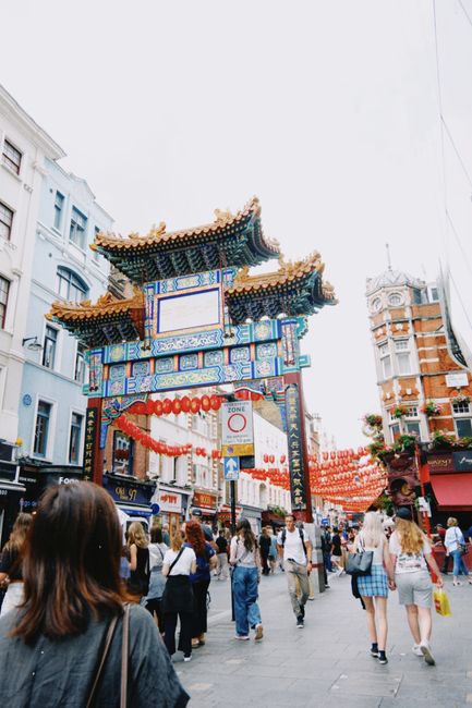 This image captures a busy street scene at Piccadilly Circus in Chinatown, WC2, with a prominent traditional Chinese-style archway featuring intricate blue, gold, and red decorative elements and a til